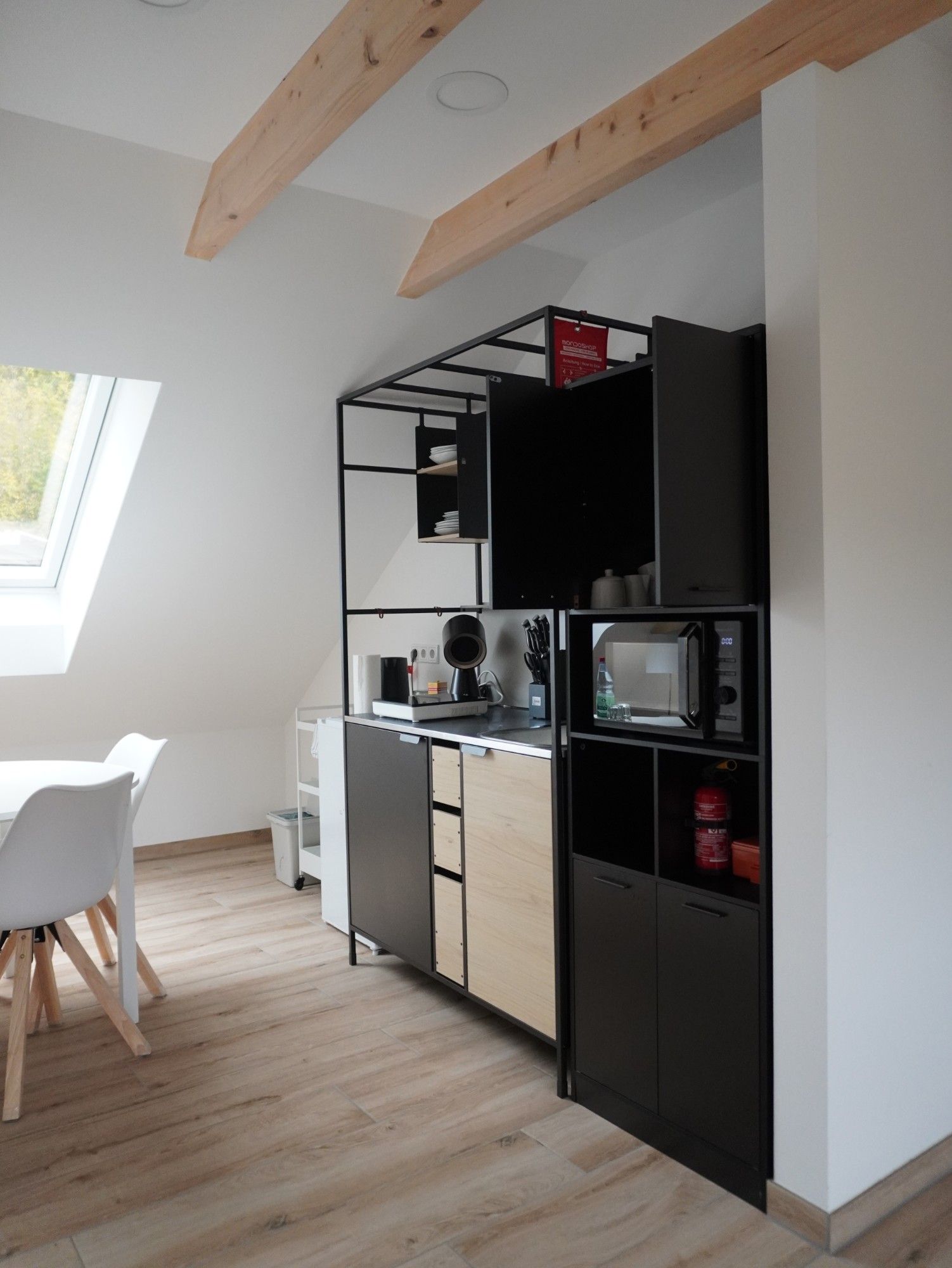 Küche im Apartment unter Dachbalken: Spüle, Wasserkocher, Messerblock, Mikrowelle und Schränke, links Dachfenster und Esstisch