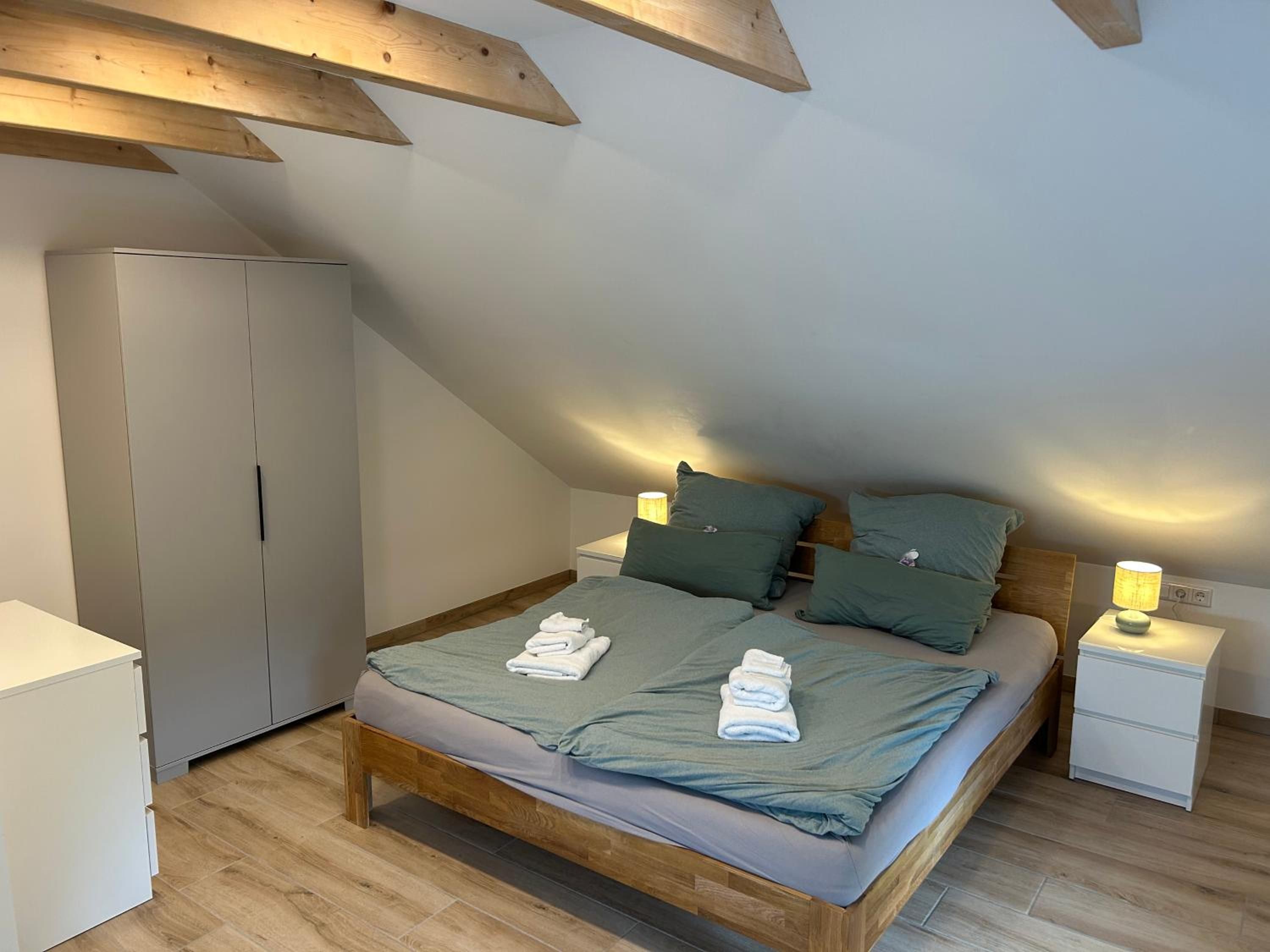 Schlafbereich im Apartment: Doppelbett aus Holz unter Dachbalken, Nachttische mit Lampen, Kleiderschrank