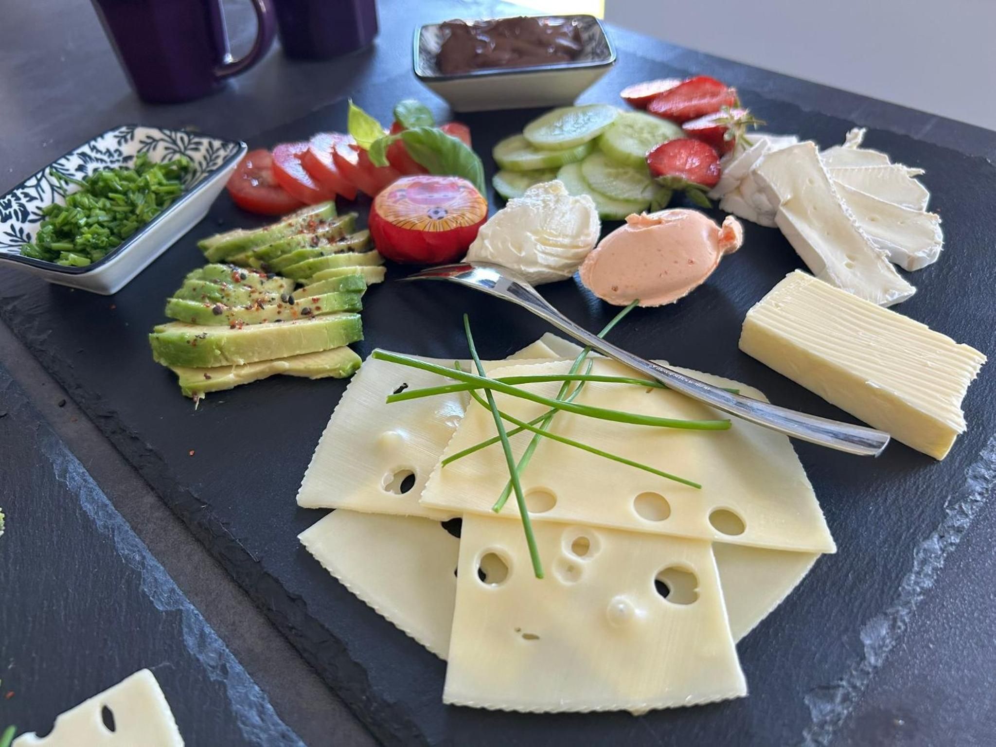 Frühstücksplatte aus Schiefer von nahem: Schnittkäse mit Schnittlauch, Camembert, Butter, Avocado, Tomaten mit Basilikum, Gurke und Erdbeeren