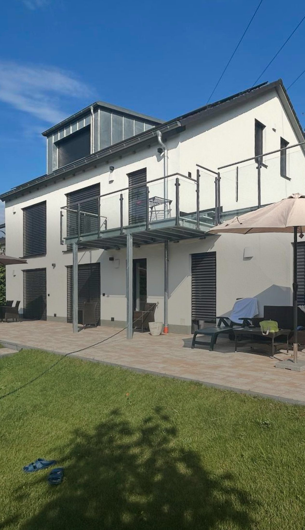 The house from the garden side: gable with dormer, balcony with glass railing, below it the terrace with seating and a hanging chair on the lawn, the forest behind